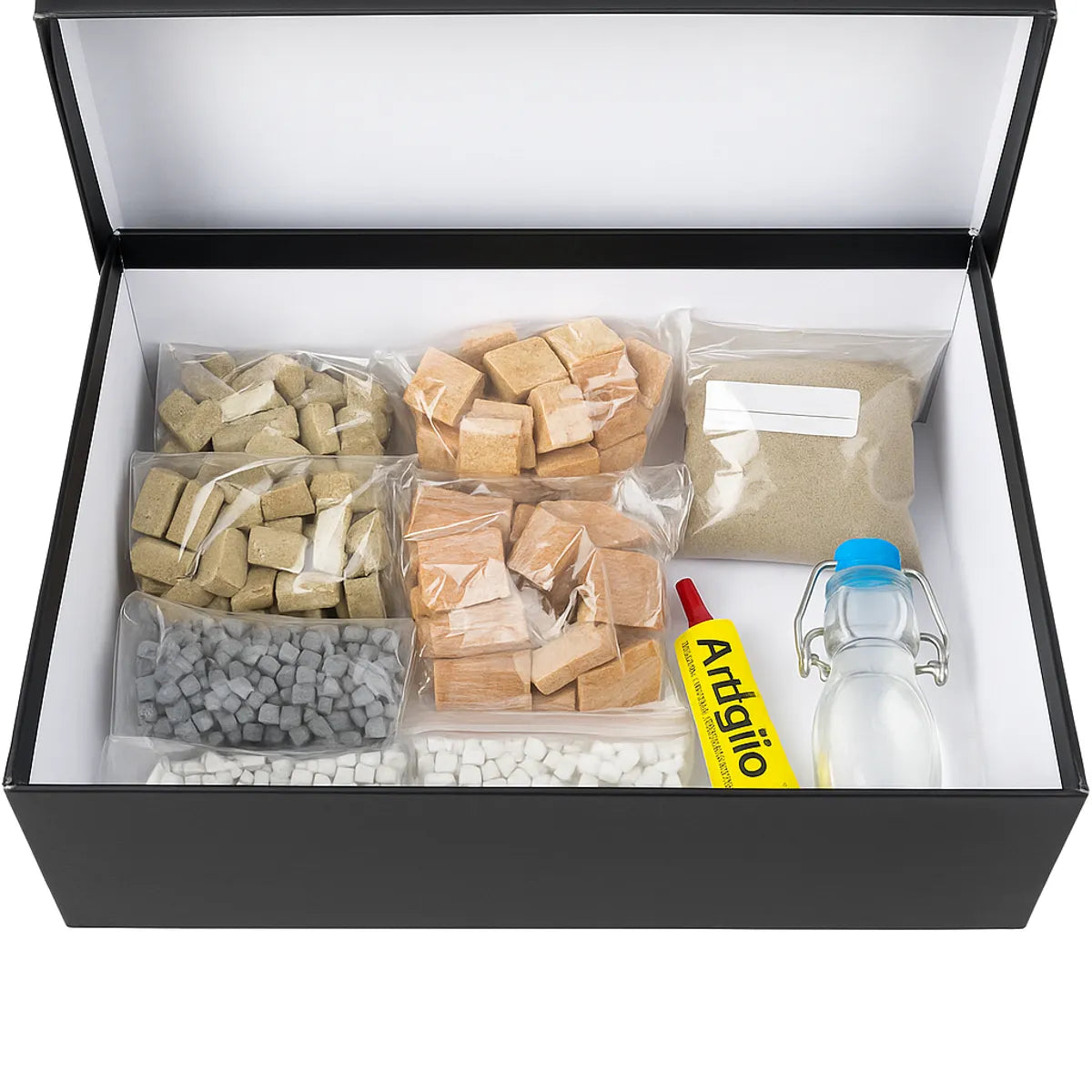Kit artistico in scatola nera con materiali per mosaico: tessere in diverse forme e colori, sabbia fine, colla Artiglio e bottiglia di vetro con tappo azzurro, disposti ordinatamente all’interno della confezione.