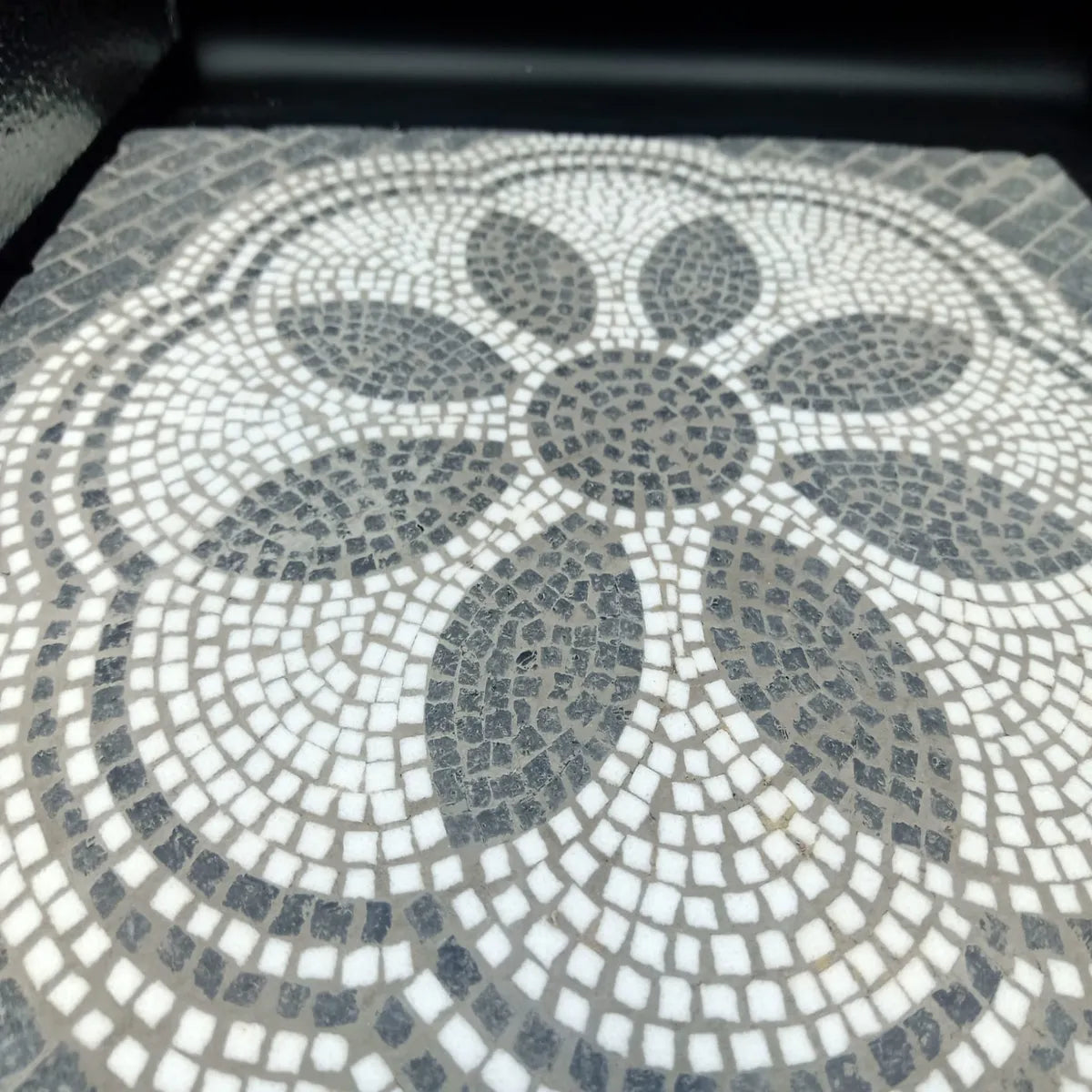 Dettaglio ravvicinato di un mosaico artigianale in pietra con motivi ad arco concentrici, tessere nere e bianche su fondo marrone, texture tridimensionale.