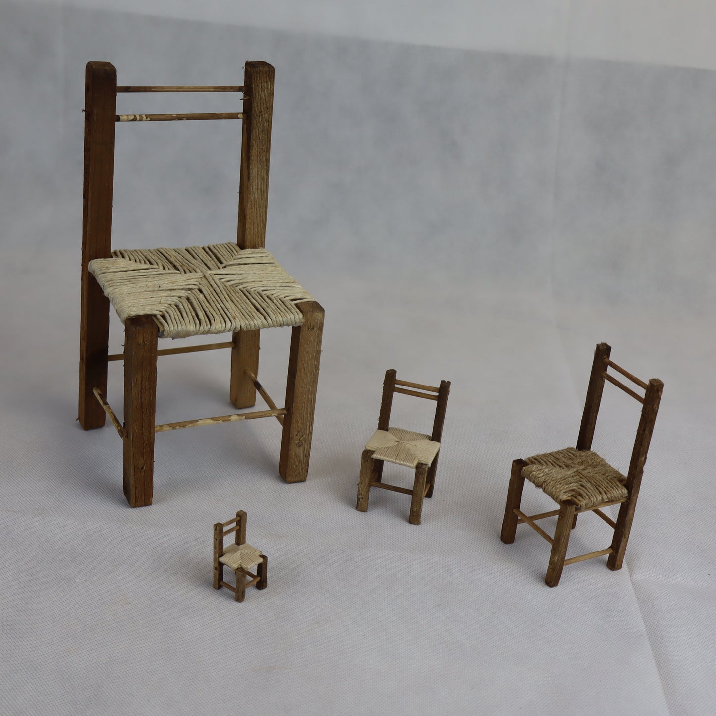 Quattro sedie rustiche in miniatura realizzate in legno grezzo, con seduta intrecciata in paglia naturale.