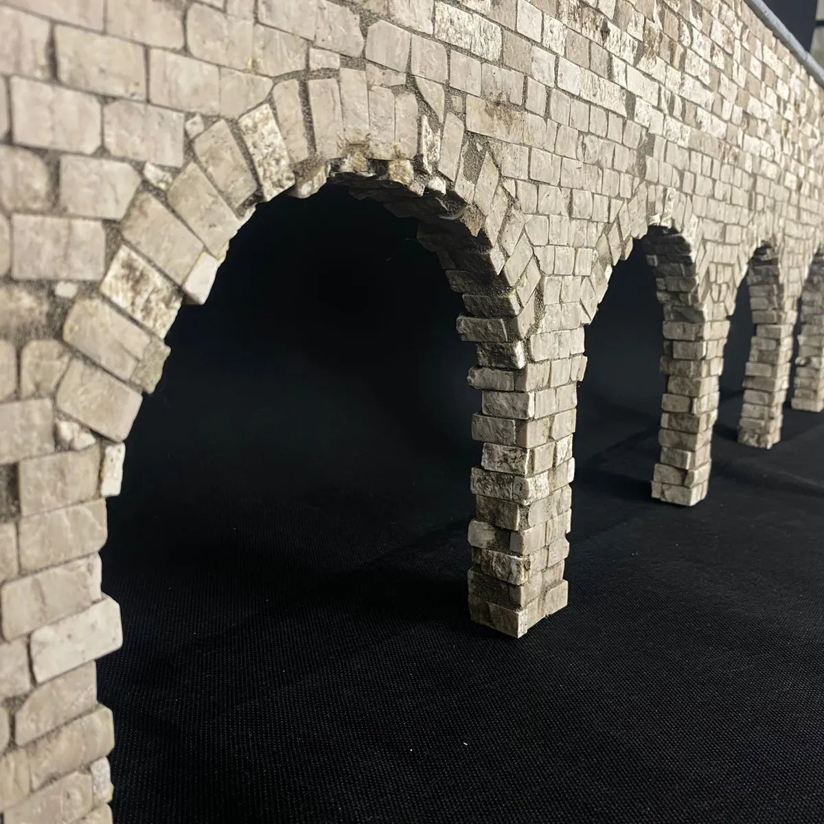 Struttura in miniatura con archi in pietra realizzati con piccoli blocchi rettangolari chiari, montata su sfondo nero per evidenziare il dettaglio artigianale delle texture.