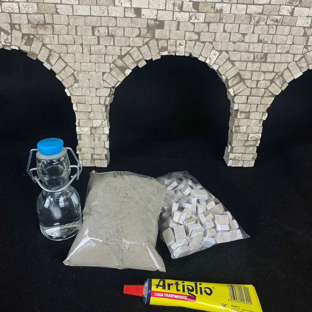 Kit “Porta Nolana” con riproduzione architettonica ad arco su sfondo bianco; include sabbia, tessere in pietra bianca, colla Artiglio e bottiglia in vetro con tappo azzurro.