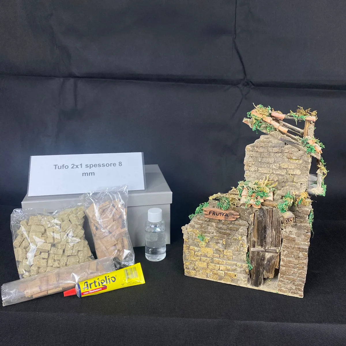 Kit completo per rudere in stile presepe: confezione con materiali accanto al rudere finito in mattoncini di tufo, porta in legno e decorazioni in muschio su sfondo nero.