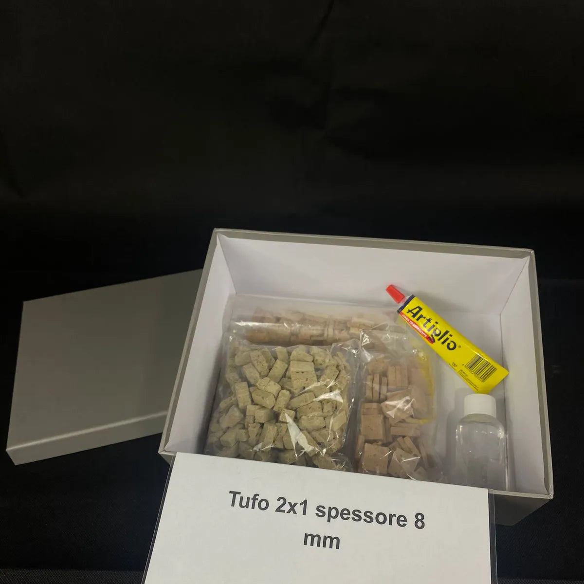 Vista dall’alto della confezione del kit “Costruisci il tuo rudere”: scatola aperta con tessere in tufo, colla Artiglio, bottiglietta e cartoncino con indicazione “Tufo 2x1 spessore 8 mm”.