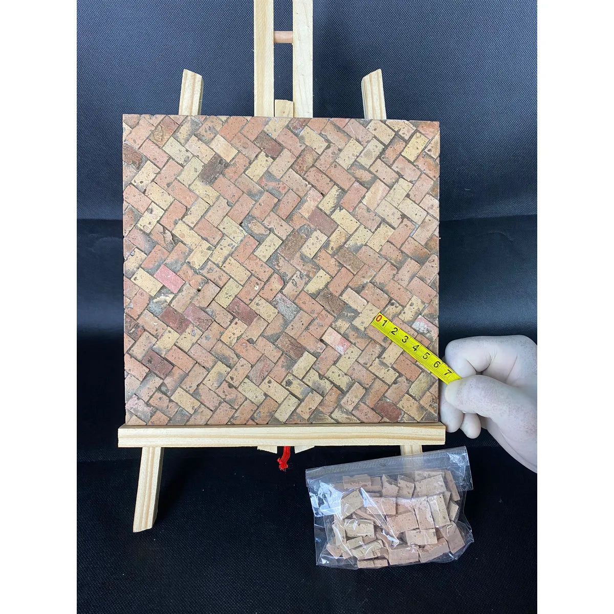 Mosaico artigianale in cotto rosato e giallo a schema spina di pesce, esposto su cavalletto e accompagnato da mattoncini confezionati.