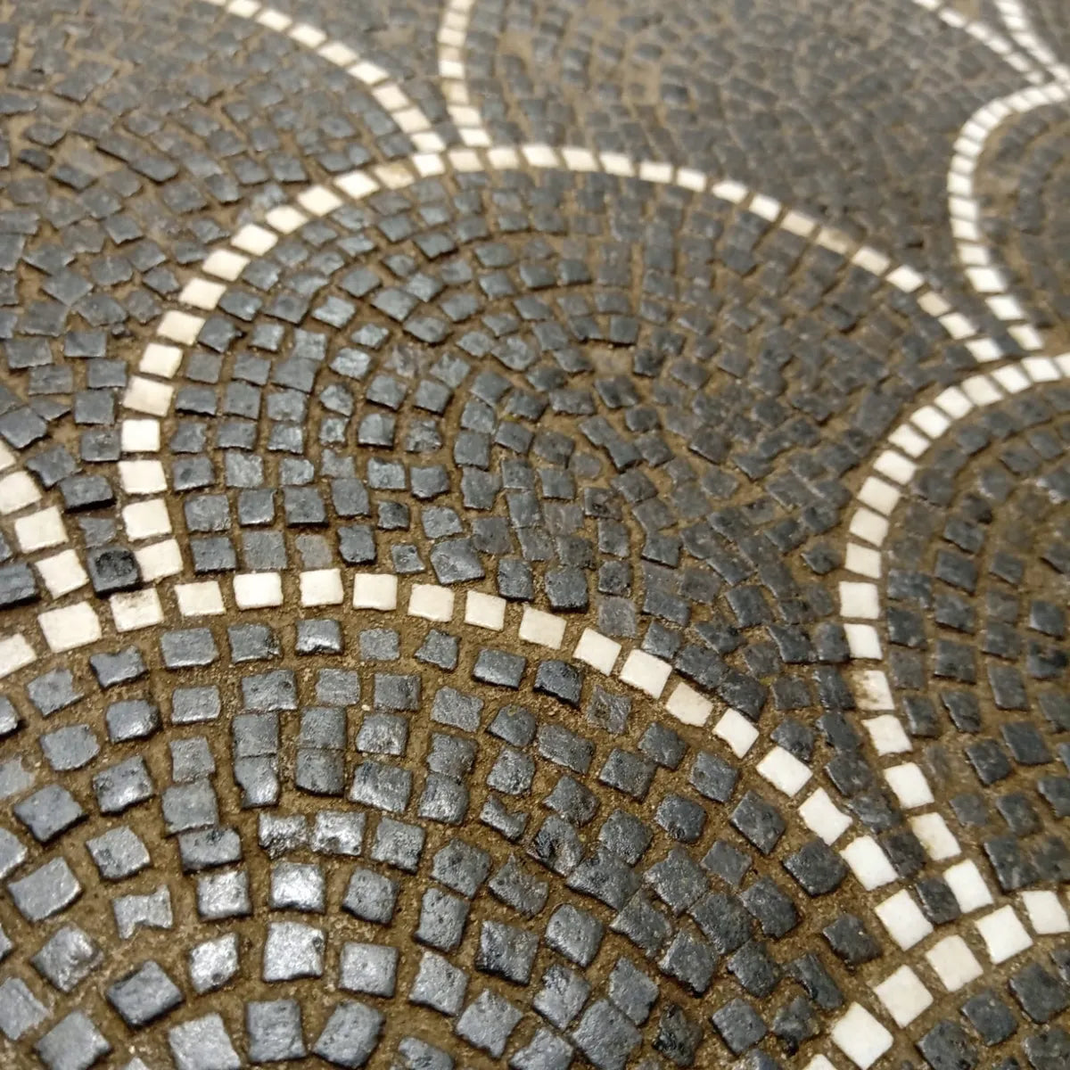 Vista ravvicinata di un mosaico in pietra con disegno ad archi sovrapposti e tessere quadrate in tonalità scura, evidenziando la precisione della lavorazione manuale.