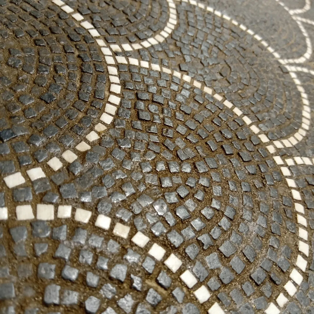 Dettaglio ravvicinato di un mosaico artigianale in pietra con motivi ad arco concentrici, tessere nere e bianche su fondo marrone, texture tridimensionale.