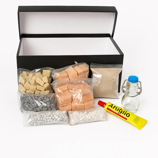 Contenuto di un kit per mosaico disposto davanti alla scatola nera: sacchetti trasparenti con tessere in pietra di varie tonalità, sabbia, tubetto di colla Artiglio e bottiglia in vetro con tappo blu.