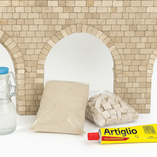 Kit per la realizzazione di archi in pietra: sabbia fine, tessere in pietra, colla Artiglio e bottiglietta in vetro disposti davanti alla struttura ad arco in muratura ricostruita.