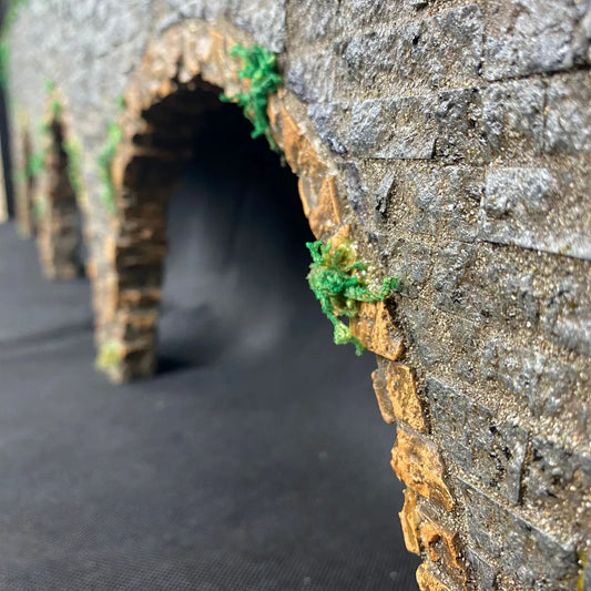Struttura in miniatura con muro in pietra e archi, realizzata artigianalmente per presepe; dettagli realistici con muschio decorativo e texture muraria effetto anticato.