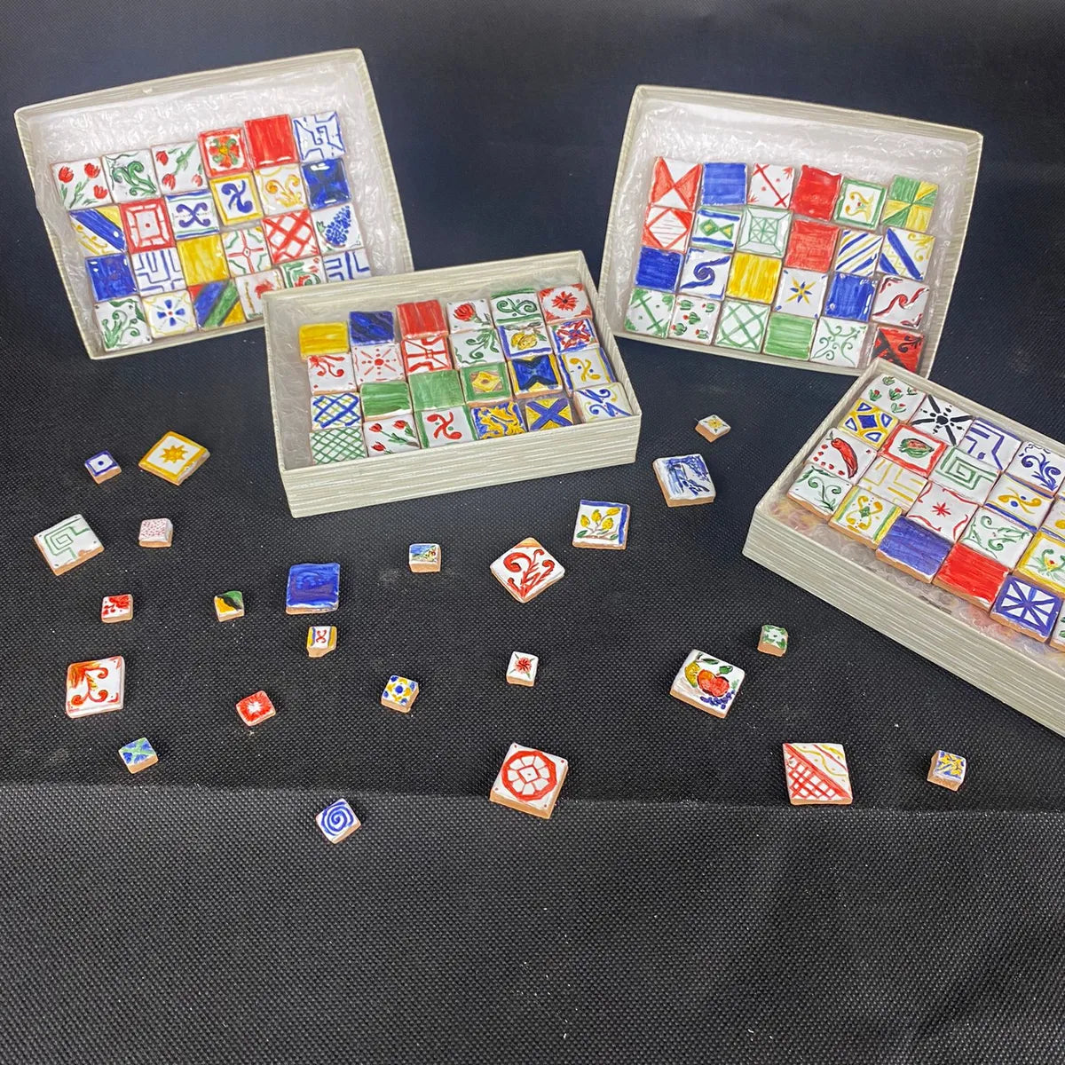 Set di piastrelle in miniatura dipinte a mano con motivi mediterranei colorati, presentate in vassoi e sparse su fondo nero per esposizione artistica.